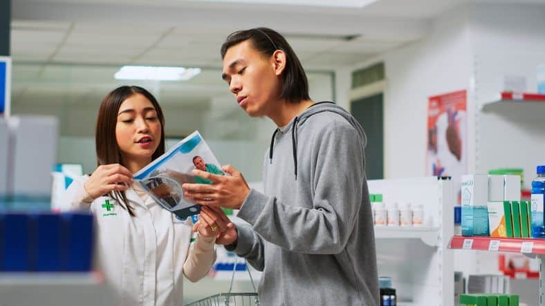 A pharmacist (or pharmacy tech) standing at a counter inside a clinic-adjacent pharmacy, handing a prescription bag or explaining medication to a patient. The vibe is trust, accessibility, and care.